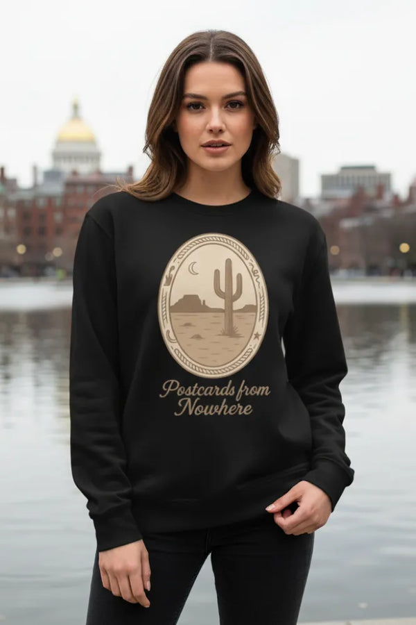 Woman wearing a black sweatshirt with a cactus design and text, standing by a body of water with buildings in the background.