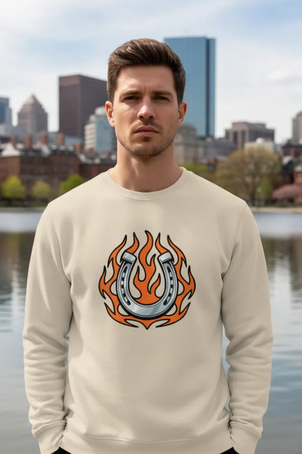 Man wearing a beige sweatshirt with a graphic design of a horseshoe and flames, standing by a waterfront with a city skyline in the background.