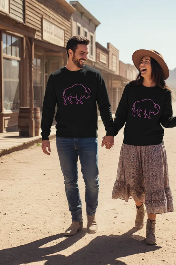 Man and woman walking hand-in-hand on a western-style street, both wearing black sweatshirts with a pink buffalo design.