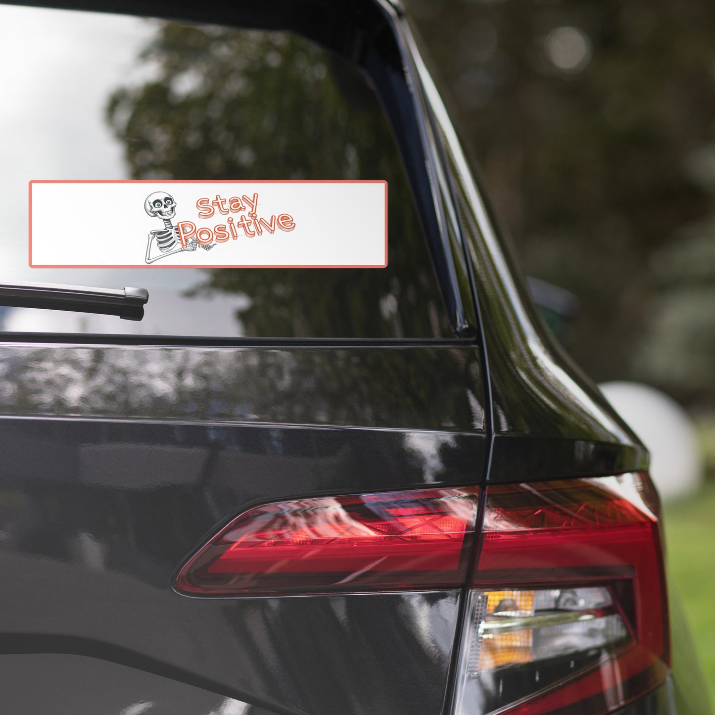 Photo of a car's rear window featuring a bumper sticker that reads 'Stay Positive' with a smiling skeleton graphic. The sticker has a white background with a light orange border, blending fun and motivation. The car is parked outdoors, with greenery reflected on the car's glossy black surface.