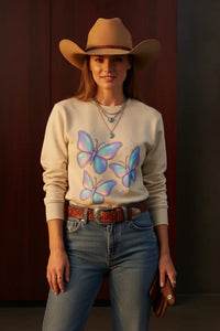 Person wearing a beige sweater with butterfly design, blue jeans, and a cowboy hat against a dark background.