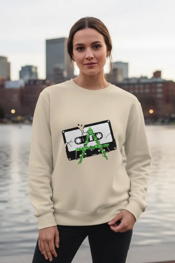 Person wearing a beige sweatshirt with a graphic design, standing by a waterfront with buildings in the background.