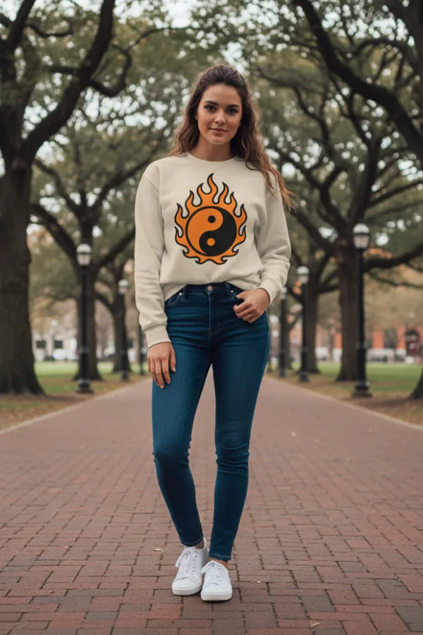 Woman wearing a beige sweatshirt with a fire and yin-yang design, standing on a brick path with trees in the background.